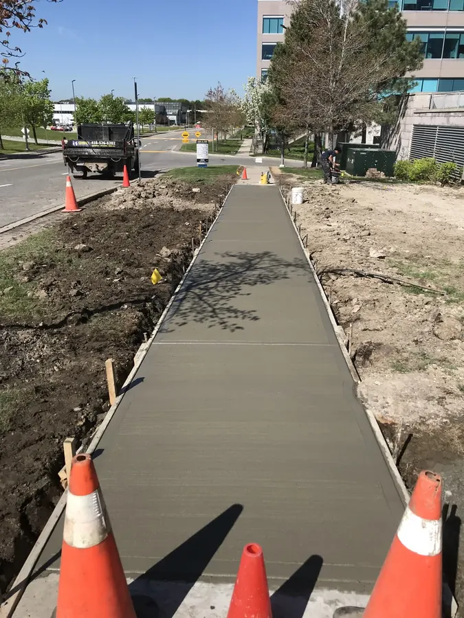 Long freshly poured broom-finished concrete walkway with timber forms, running through an office park