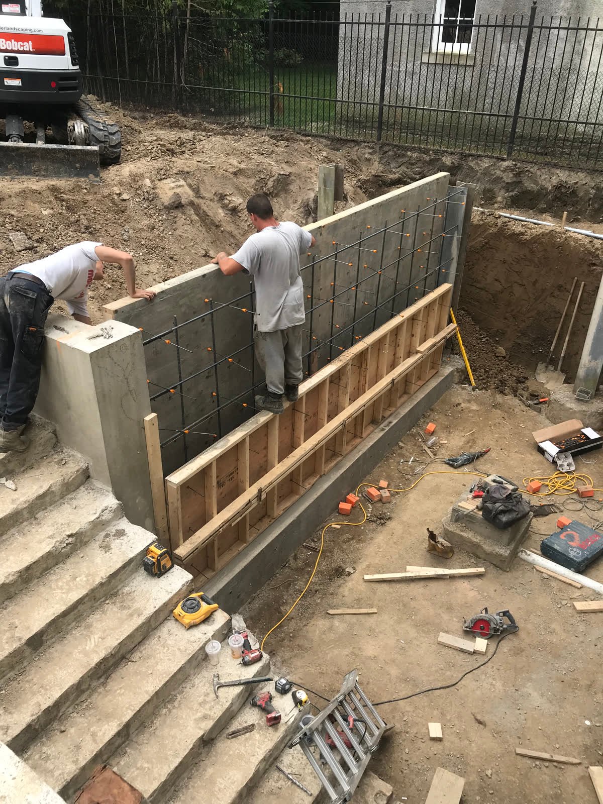 Two workers setting timber forms against a tied rebar mat for a poured concrete retaining wall